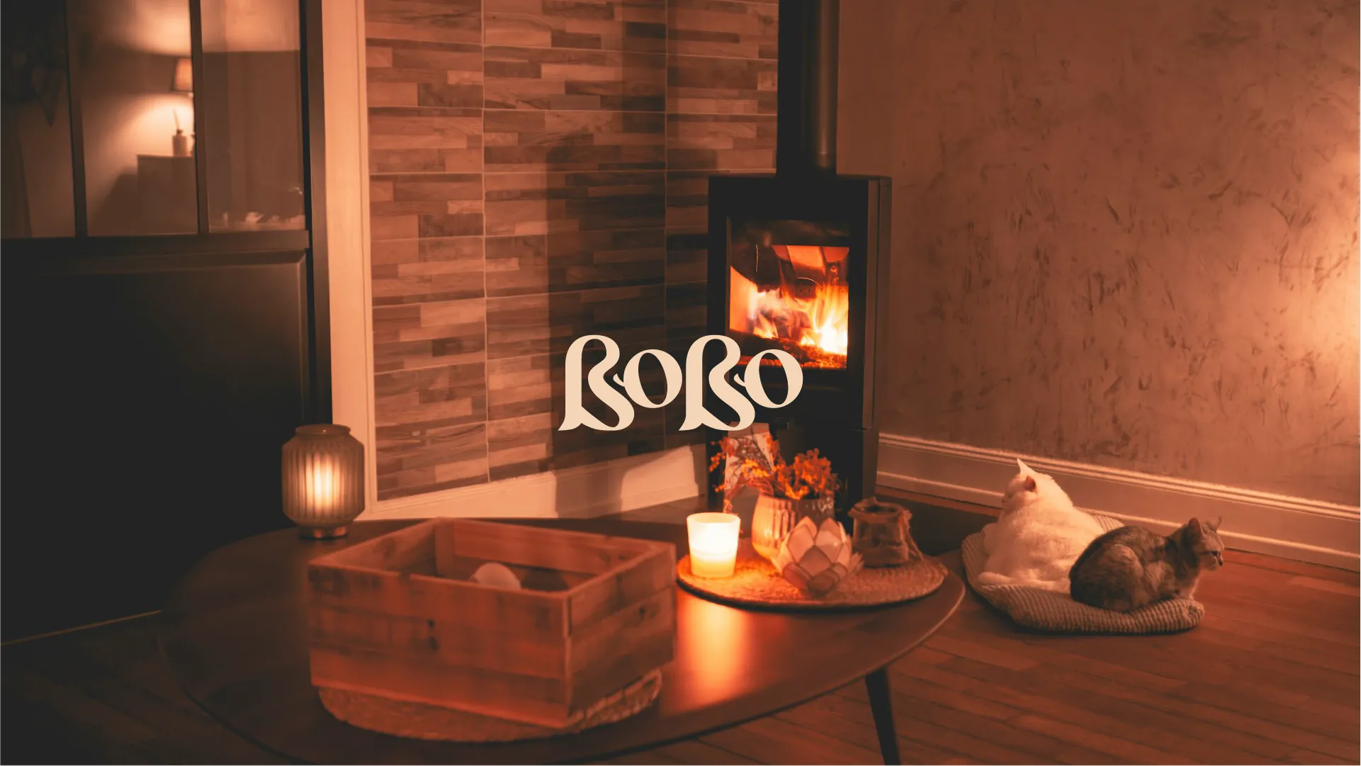 The Bobo wordmark over a warm, candle-lit living room at night, with a wood-burning stove and two cats.